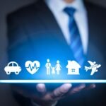 Imagem ao fundo, desfocado, de um homem vestido com terno e gravada, imagem focada na mão desse homem segurando um tablet, acima desse tablet ícones que remetem ao seguro de veículo com ícone de carro, vida com ícone de um coração, familiar com ícone de um boneco masculino, um boneco de criança e uma boneca feminina, residencial com ícone de uma casa, e viagem com ícone de um avião.