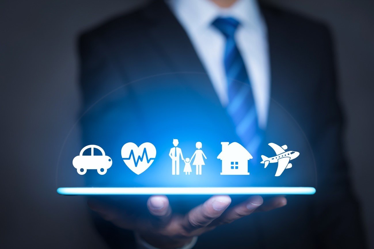 Imagem ao fundo, desfocado, de um homem vestido com terno e gravada, imagem focada na mão desse homem segurando um tablet, acima desse tablet ícones que remetem ao seguro de veículo com ícone de carro, vida com ícone de um coração, familiar com ícone de um boneco masculino, um boneco de criança e uma boneca feminina, residencial com ícone de uma casa, e viagem com ícone de um avião.